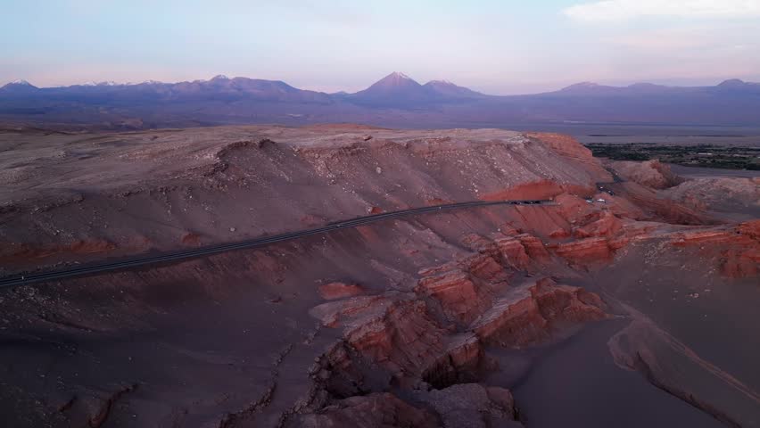 Ascending drone shot rising over a desert road cutting through red rock formations revealing the Atacama oasis and snow capped volcanic peaks at dusk. Valle de la Luna, San Pedro de Atacama, Chile