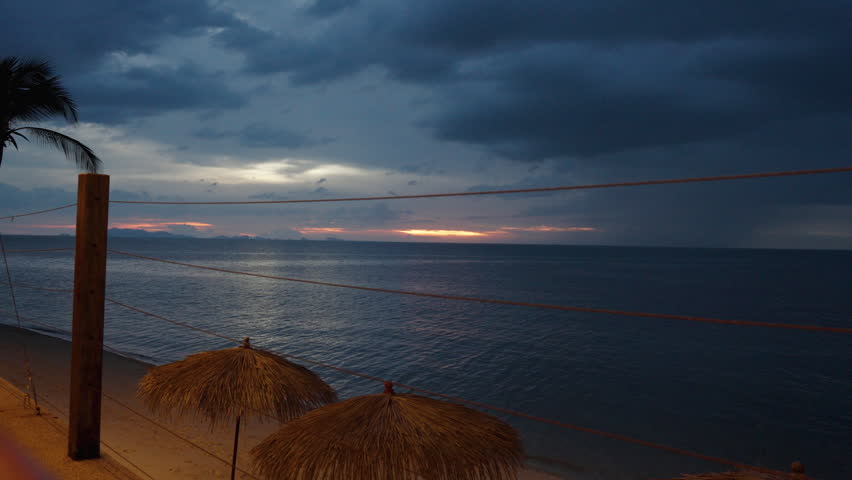 Yellow Cocktail with Orchid Decoration Stands on Bar Table as Camera Shifts Focus from Distant Sunset Over Ocean to Drink in Foreground, Tropical Evening Relaxation Concept.