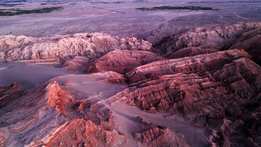 High altitude drone shot over dramatic red and purple eroded rock formations glowing in warm dusk light with the vast Atacama desert plateau stretching beyond. Valle de la Luna, San Pedro de Atacama,