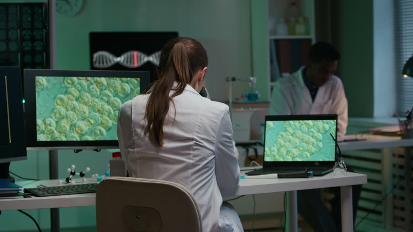 Back view of young female scientist in white coat decoding genomic sequences with microscope while sitting at table with molecular system on computer screen in laboratory