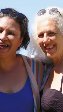Vertical video: Smiling pair leaning in at beach as camera starts, wearing swimsuits and sunglasses. Friends, outdoor, sunlit, swimwear, shades, poolside, vertical video
