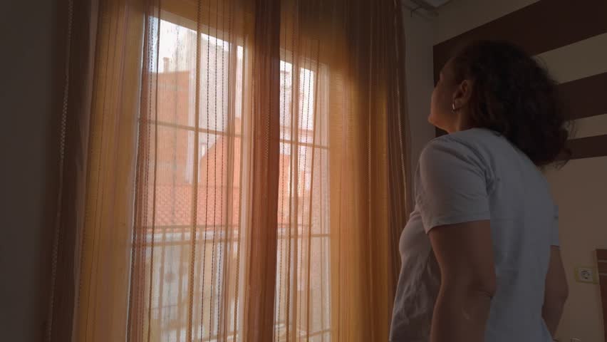 A woman standing by a window, gently opening curtains to let in the morning sunlight.