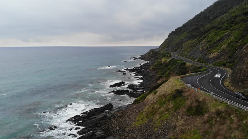 Cinematic aerial drone footage tracking forward along the iconic Great Ocean Road in Victoria, Australia. The road runs along the right of the frame while the vivid blue coastal waters of the Southern Ocean fill the left, with the rugged coastline stretching ahead into the distance. Shot on a cloudy midday, the overcast conditions add a dramatic and moody quality to this sweeping coastal perspective.
