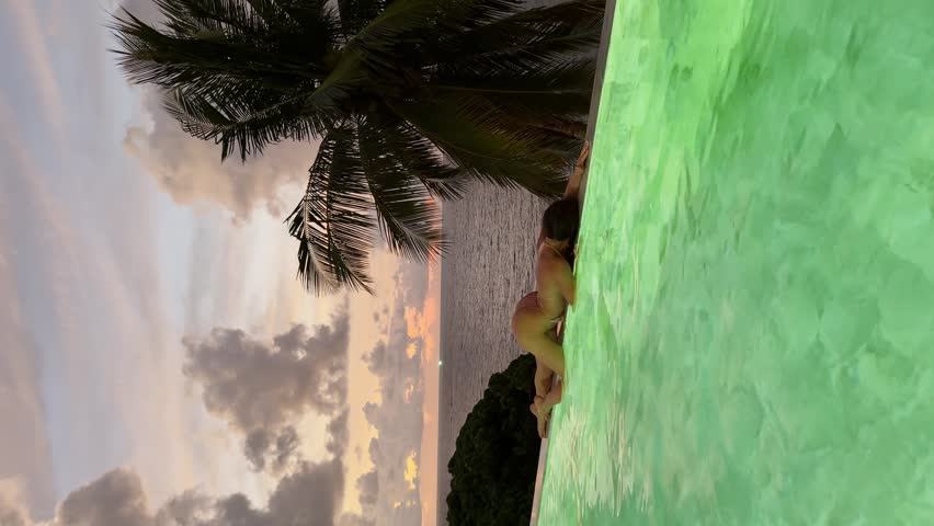 Thailand, Phuket, A girl is having fun on the edge of a swimming pool in a luxury villa at sunset, with a coconut palm and the sea in the background