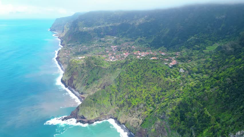 Dramatic aerial view of a verdant Madeiran valley, revealing a Boaventura charming village nestled amidst steep, cloud-kissed mountains and ancient terraced fields.