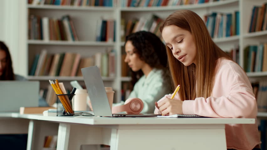 Young Girl Writing Notes at a Study Table in a Library With Other Students Nearby