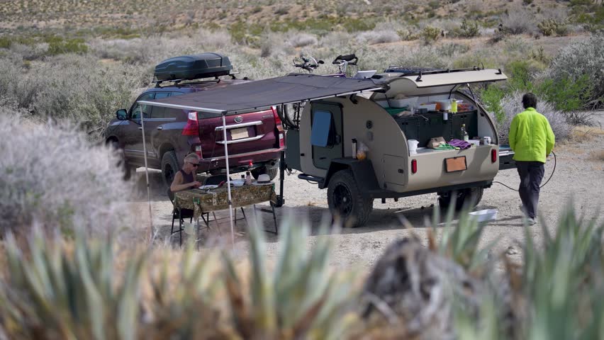A mature woman and man enjoy a meal beside their teardrop camper trailer in Plum Canyon, California while overlanding in the desert.