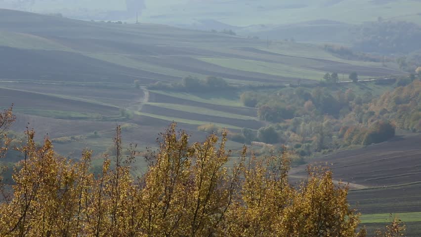 A breathtaking, hazy view of rolling hills and mountains in autumn. Terraced fields and scattered trees create a serene, expansive natural landscape.