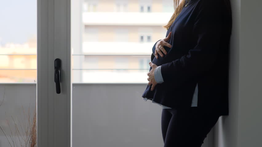 Side profile of pregnant woman in dark blazer standing beside a large window, hands cradling bump, city building visible outside, natural light