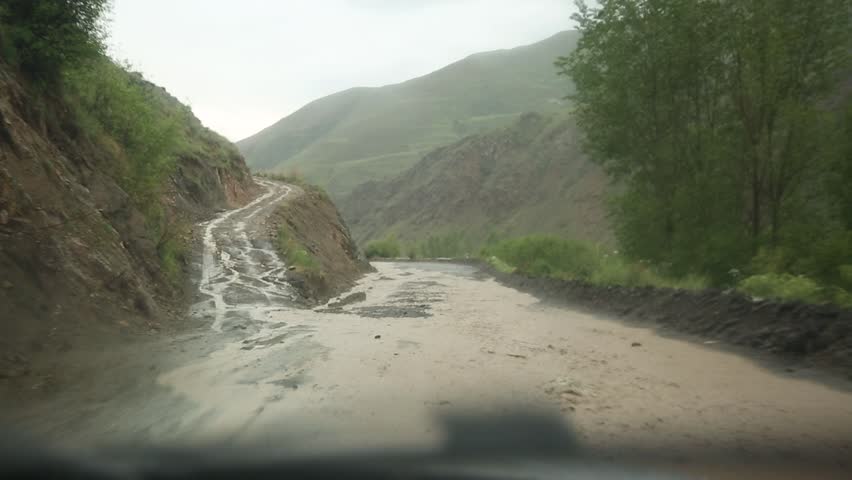 A road runs through a lush valley, partially submerged by a muddy, fast-flowing river. The water has overflowed its banks due to heavy rainfall, creating a challenging and hazardous driving condition amidst the verdant, hilly landscape.