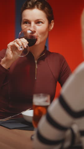 Close up of brunette woman in burgundy zip top holding a red wine glass and speaking expressively, beer glass and bowl visible in foreground, vivid red wall behind