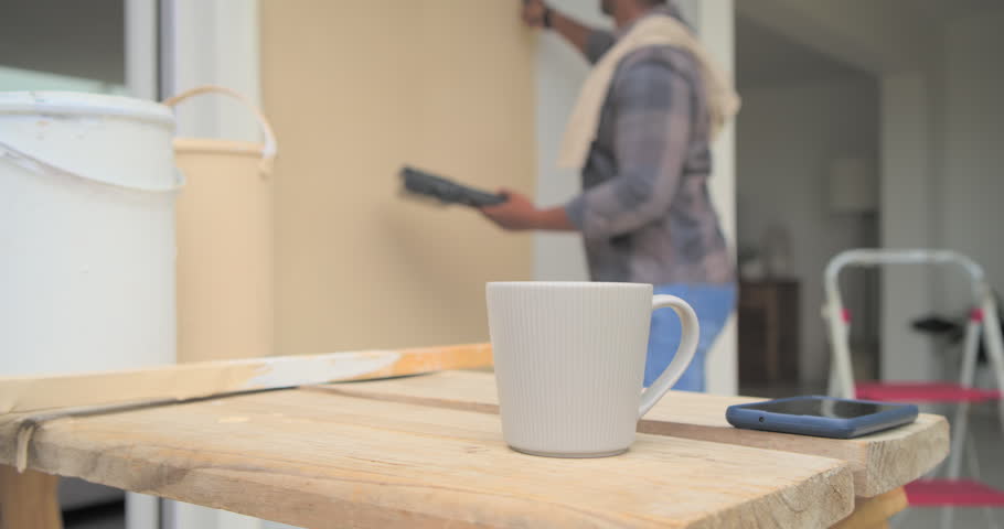 African male placing paint tray on bench, wiping hands, taking mug, phone, checking wall at doorway. Exterior, porch, renovation, craftsman, roller, beige, plaid