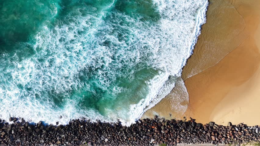 Aerial drone downward video showing waves rolling onto Nobbys Breakwall at the entrance to Newcastle Harbour in New South Wales, Australia. Ocean swell creates whitewater patterns as it meets the long rock structure extending into the Pacific Ocean. Scenic coastal perspective captured from above highlighting natural water movement and maritime infrastructure along the rugged shoreline.