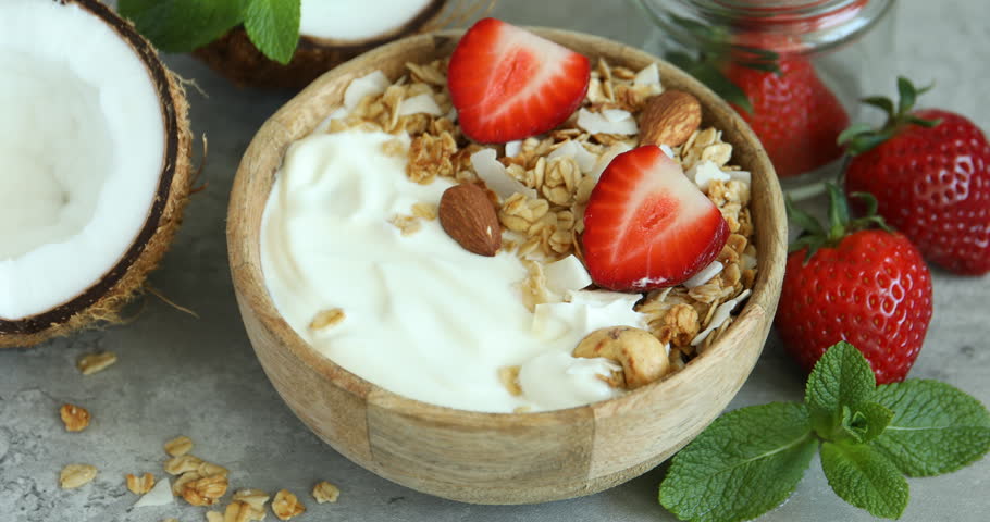 Woman taking delicious coconut yogurt with granola, strawberry and nuts from bowl with spoon at grey textured table, closeup