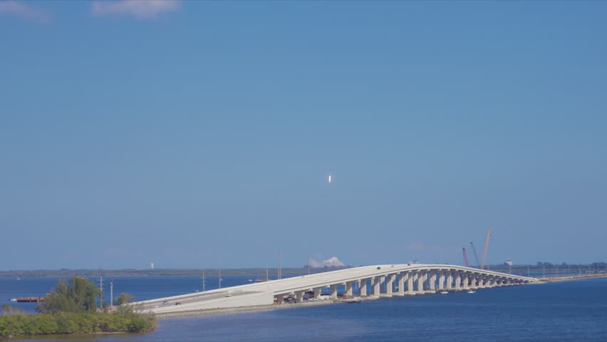 Rocket ascending into clear blue sky with exhaust trail visible above long concrete bridge spanning calm blue water. Cars drive across as cranes work in background on sunny day