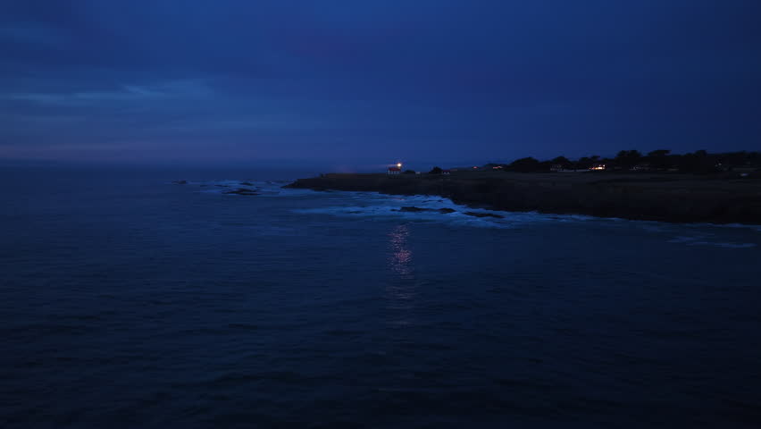 Night aerial view of Point Cabrillo Lighthouse in Mendocino, California, USA. Waves crash against rocky coastline as beacon light glows against deep blue twilight sky with distant buildings visible