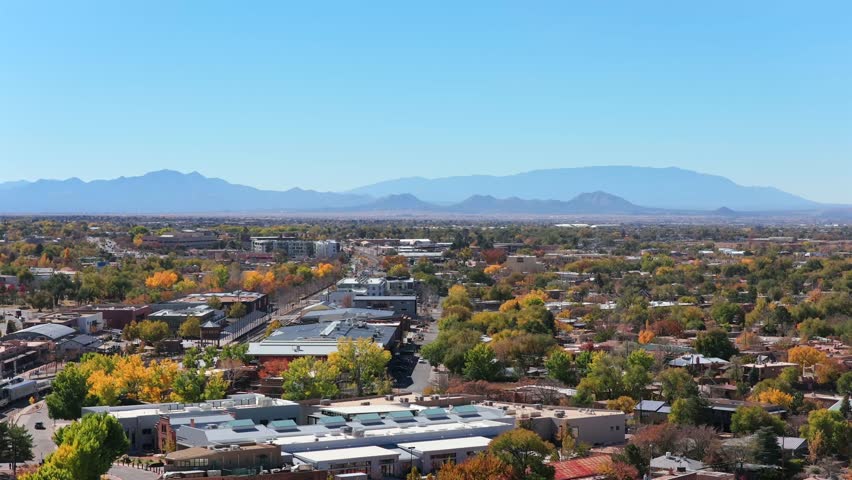 A rising drone shot captures the Santa Fe cityscape with the historic railyard area on the left. The footage highlights unique Southwest architecture and urban layout under a clear blue sky.