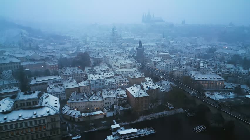 Winter morning aerial view of snowy Prague rooftops with misty castle panorama and historical bridge.