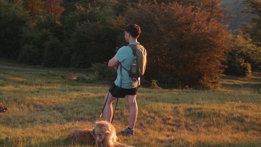 A man and a golden retriever pause together on a grassy hill, both looking forward into the light. Captured in 4K at dusk.