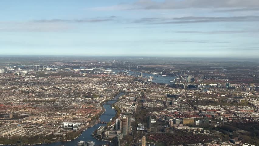 High-altitude city view over Amsterdam showing dense urban neighborhoods, waterways, and harbor areas under a pale blue sky.