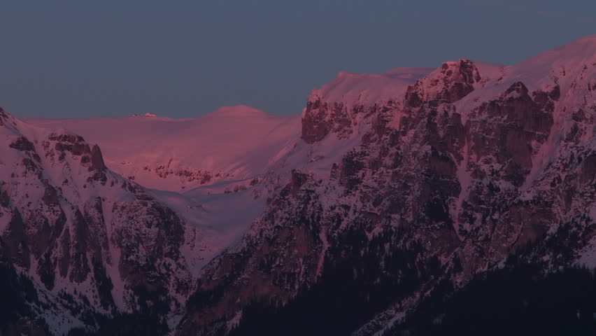 Immersive deep pink alpenglow on the steep snow capped peaks of Gaura valley and Scara in the Bucegi Mountains, high altitude aerial pan left super telephoto cinematic 4K