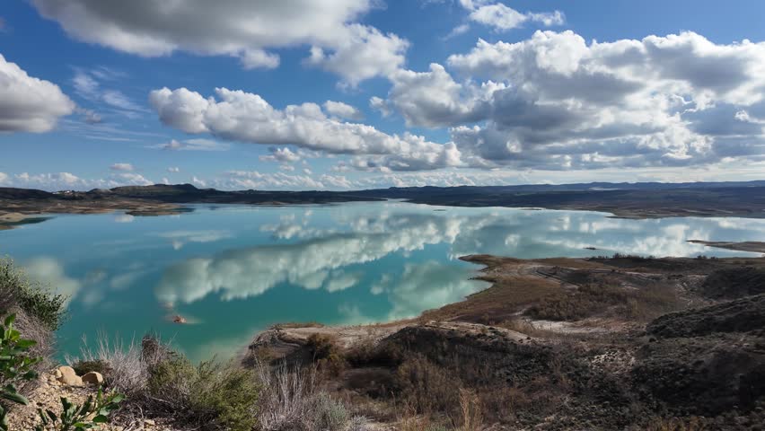 Scenic lake with calm water reflecting clouds and blue sky, peaceful natural landscape with mountains and shoreline, tranquil outdoor environment