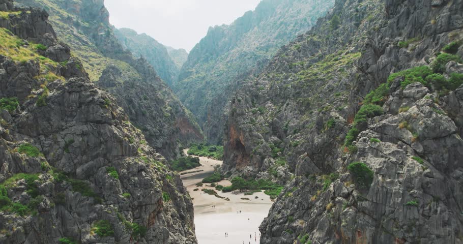 Drone shot captures the impressive Torrent de Pareis canyon winding through rugged mountains in Mallorca, Spain. Discover authentic natural beauty and geological wonder with people exploring its dry riverbed.