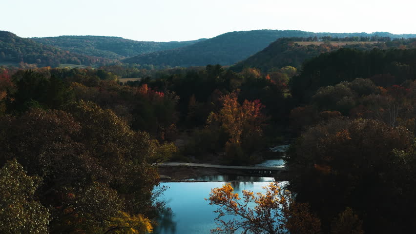 Calm river reflecting colorful autumn forest hills, Natural Dam Arkansas Ozark Mountains, drone pullback