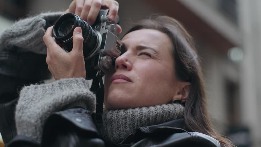 A focused woman photographer takes pictures with a retro camera on a city street. She looks through the viewfinder and checks her camera.