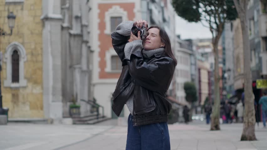 A young female tourist takes pictures of city architecture with a retro camera. She smiles while exploring the historic streets on her vacation.