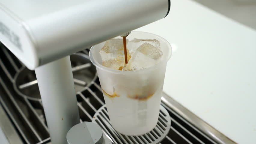 rich espresso being poured into a transparent cup of cold milk and ice cubes to create an iced coffee latte. Professional barista preparation in a clean, minimalist cafe setting.