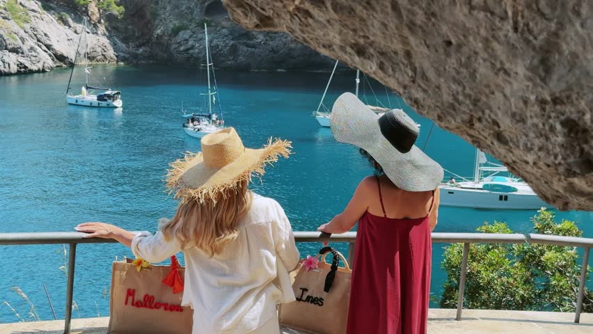 Two women with straw hats look out over the beautiful blue waters of Sa Calobra, Mallorca, with yachts and boats anchored in the scenic bay. Capturing a genuine travel experience in Spain.