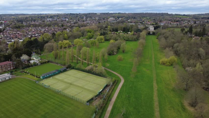 High angle aerial view of vibrant green sports fields and public parkland in Canons Park, Stanmore. Cinematic drone shot showcasing football pitches, cricket grounds, and recreational areas in a suburban London community
