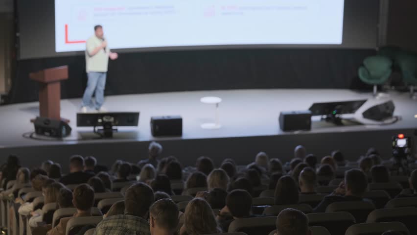 Conference speaker presenting on stage, large audience watching in dim auditorium, professional event atmosphere, live presentation and networking opportunity.