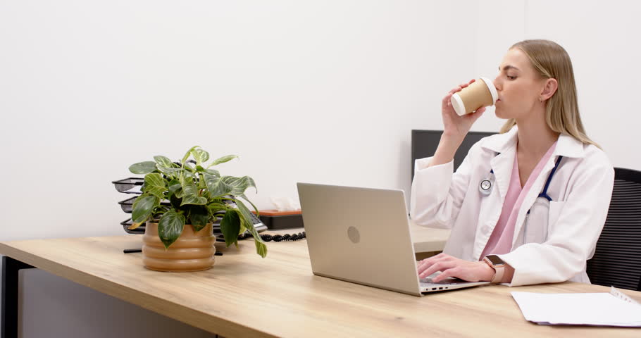 Adult female doctor typing on laptop, lifting cup and sipping while working at clinic desk. Clinician, office, workstation, productivity, focus, minimal, modern