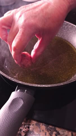 Meatball being dropped into hot olive oil frying pan by hand with steam rising. Mediterranean cooking.