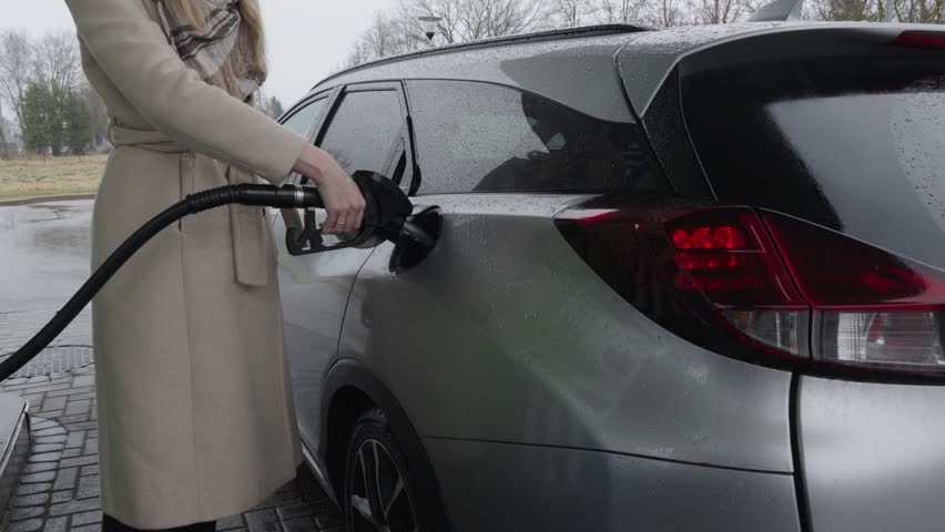 Person fills gas tank with expensive gas at station on a rainy day while wearing a coat