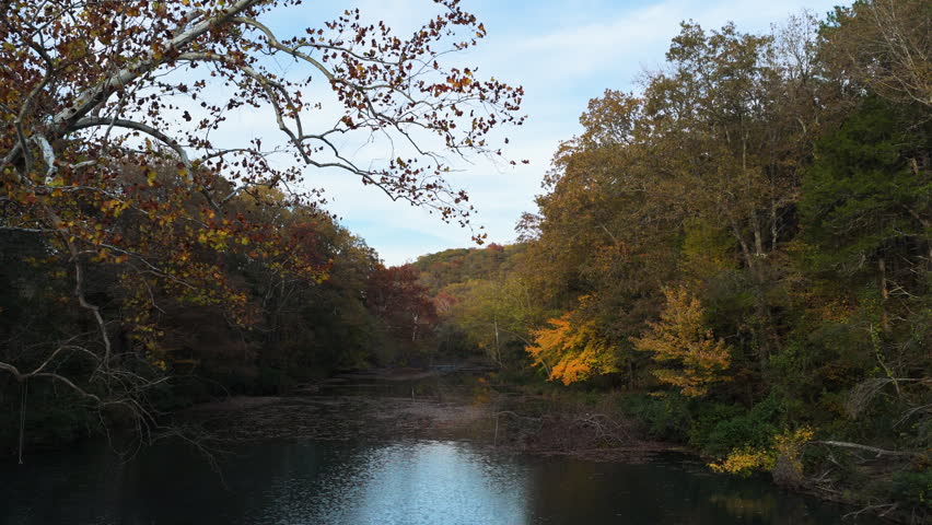 Calm forest river reflecting autumn foliage in Ozark woodland near Natural Dam Arkansas, drone medium pullback with sunset glow