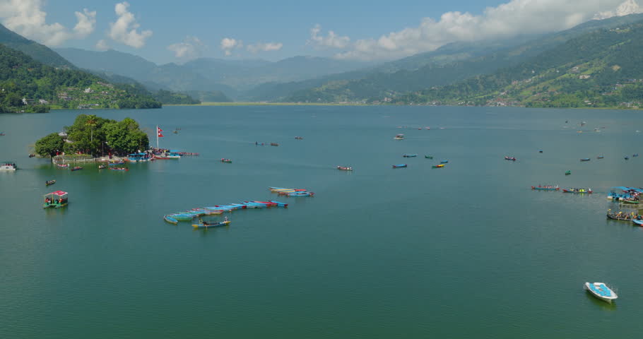 Aerial shot of Tal Barahi Temple n Phewa Lake Nepal with colorful boats floating on blue water and Nepal flag visible under a cloudy sky in Pokhara.