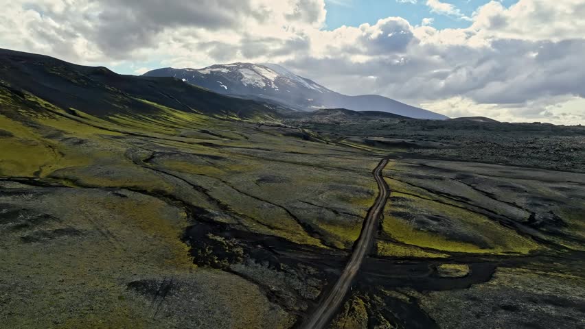 Aerial view of a winding road crossing mossy lava flats toward a snow-capped volcano, with dramatic clouds and sun rays over a vast remote highland landscape.
