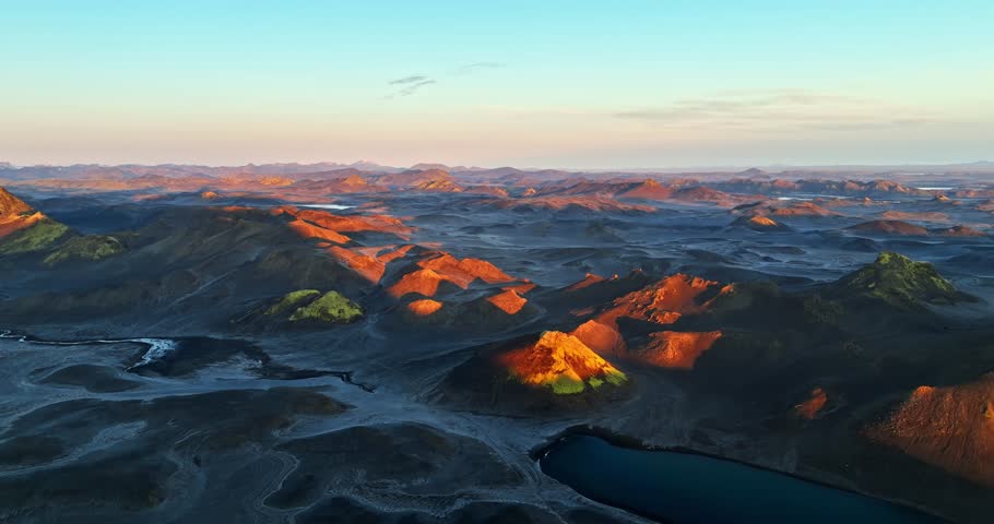 Aerial view of volcanic highlands glowing in low sun, with orange-lit ridges, dark lava plains, winding riverbeds, and a deep blue crater lake across a vast remote landscape.