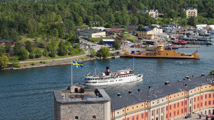 Swedish flag flying proudly on the tower of historic stone Vaxholm Fortress. Aerial view of Vaxholm city, Vaxholm fortress, passenger steamship and harbor with various sea vessels. Vaxholm city is located on the islands of Vaxån and Kullo in Stockholm archipelago, Sweden.