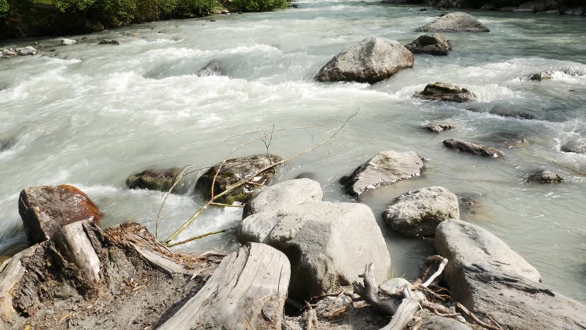 The waters of an impetuous torrent in a mountain environment, in the village of La Thuile, Aosta Valley, Italy. Rocks in the foreground. Forest on the background.