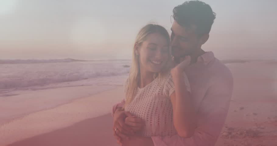 Man approaching wrapping woman behind, leaning smiling toward horizon, waves and lens flare romance. Beach, sunset, couple, embrace, warm, glow, sea