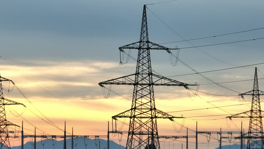 Power lines and electrical transmission towers stand tall in the rural area as the sun sets behind them. The sky shows vibrant colors.