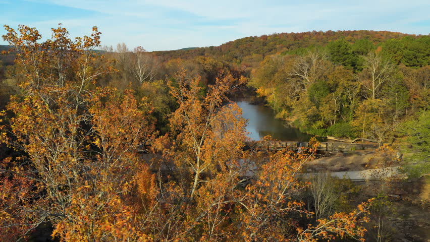 Forest river flowing beside autumn trees in Ozark Mountains Natural Dam Arkansas landscape, drone tracking pan right reveal