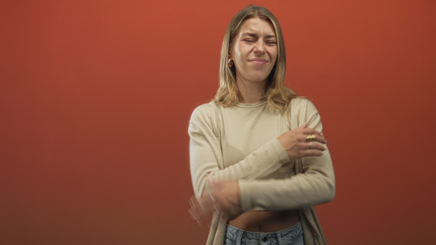 Young woman hugging arms across chest, showing bellybutton in beige crop top and jeans against orange studio backdrop; self care serenity.