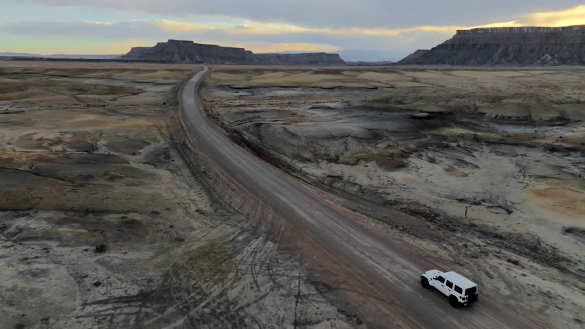 Aerial view of a white SUV driving along a dirt road through the desert landscape near Factory Butte in Wayne County, Utah, United States.