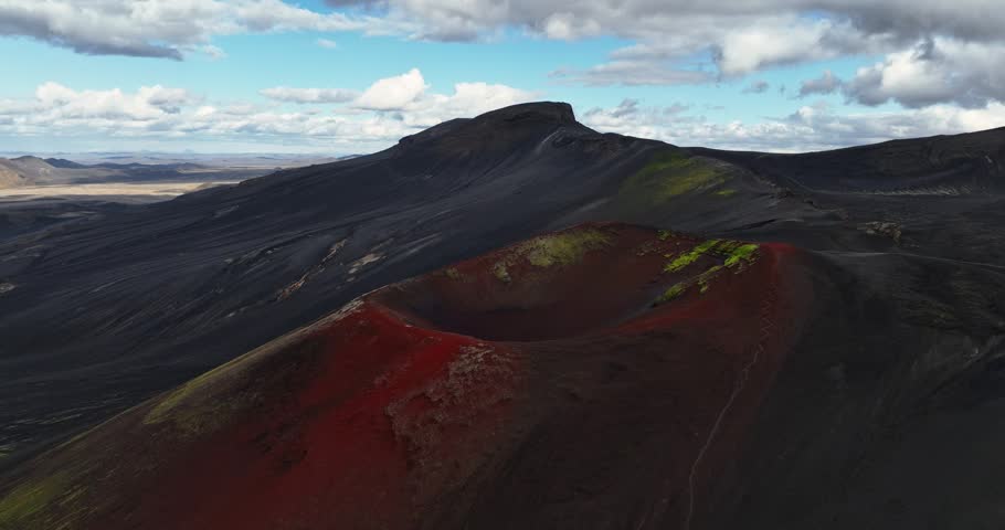 Aerial view of a vivid red volcanic crater rising from dark ash slopes, with green moss patches and rugged highland mountains under a dramatic cloudy sky.
