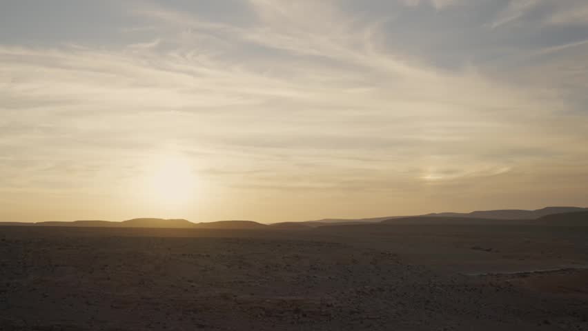 A serene desert landscape on a clear day at sunset or sunrise, sun setting behind distant hills. A peaceful and calm natural environment of blue skies with soft clouds above a wilderness land of dunes